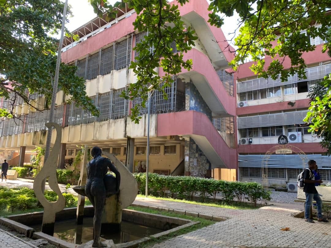 Modern academic building with pink facade, outdoor stairs, lush trees, and a sculptural fountain.