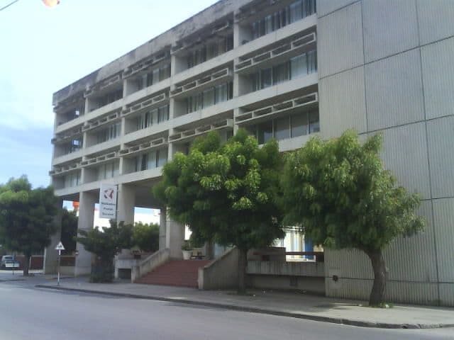 Modern concrete building with shaded balconies and green trees along a street.