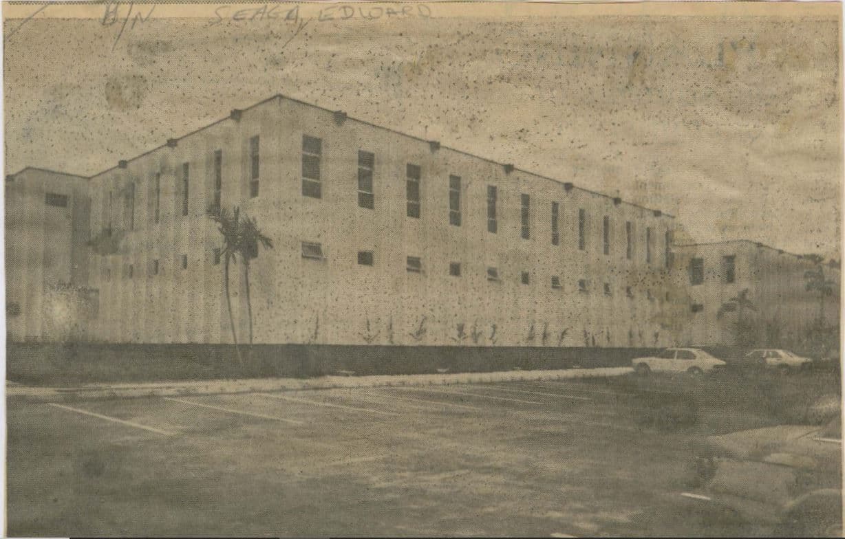 Exterior view of a long, two-story, light-colored building with many windows and a fenced perimeter.