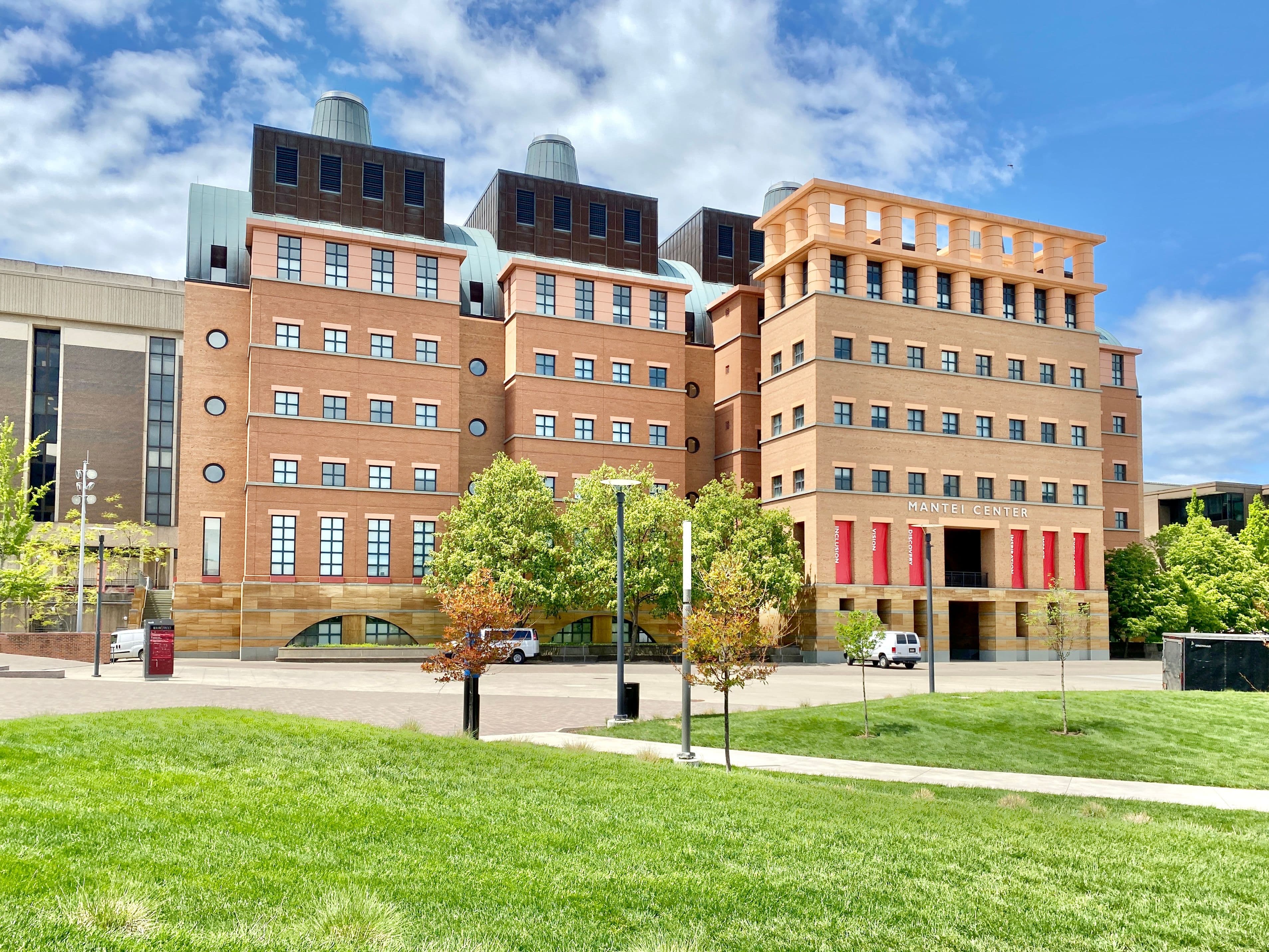 A multi-story brick building with large windows and distinctive red architectural elements stands under a blue sky.