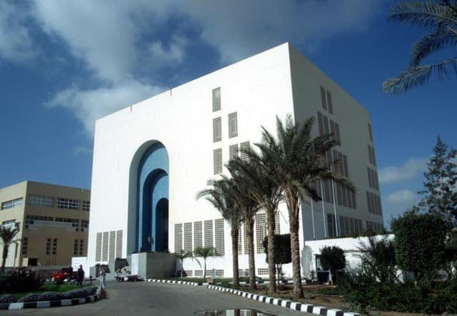 Modern white building with prominent blue arched entrances and tall palm trees under a blue sky.