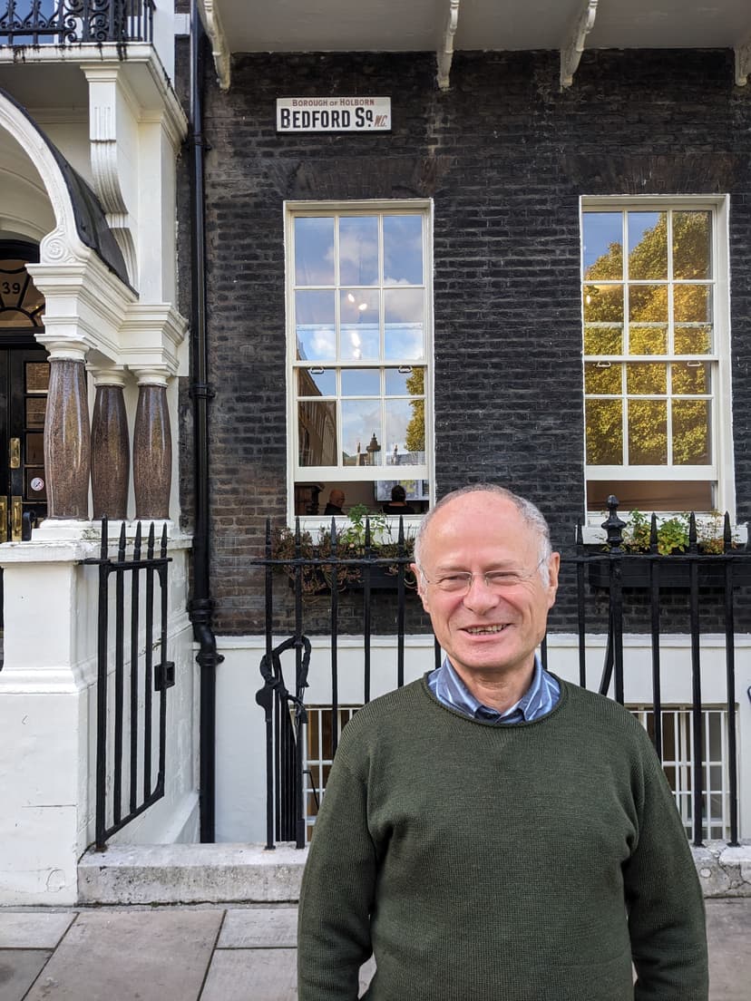 Elderly man with a balding head and a friendly smile stands outside a building.