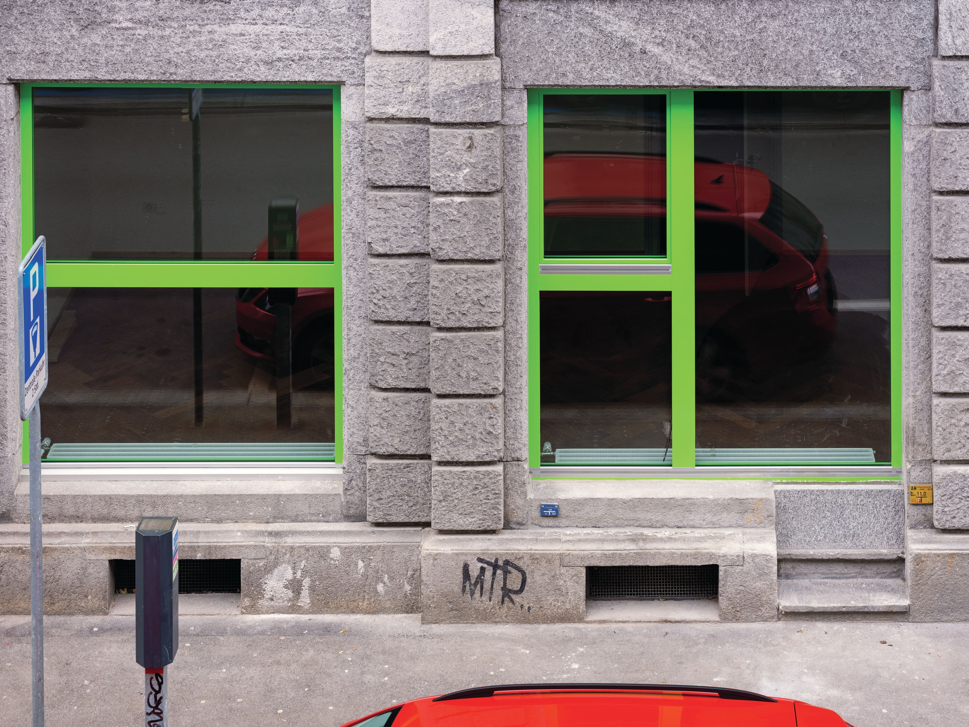 A stone building facade with bright green window frames. The top of a bright red car is visible in front of the building.