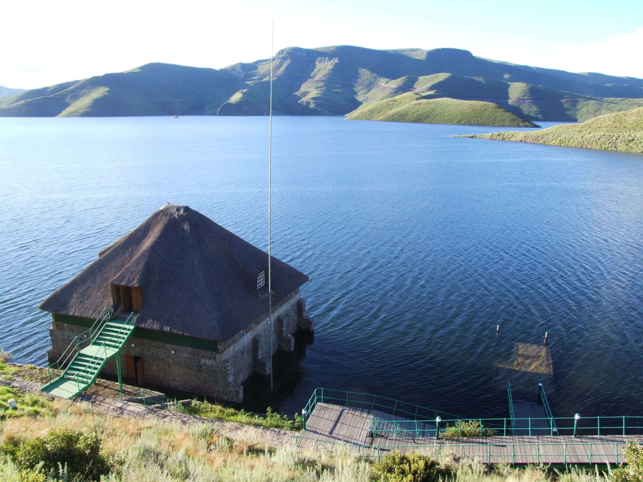 A stone building with a dark roof sits by a blue lake with distant mountains.