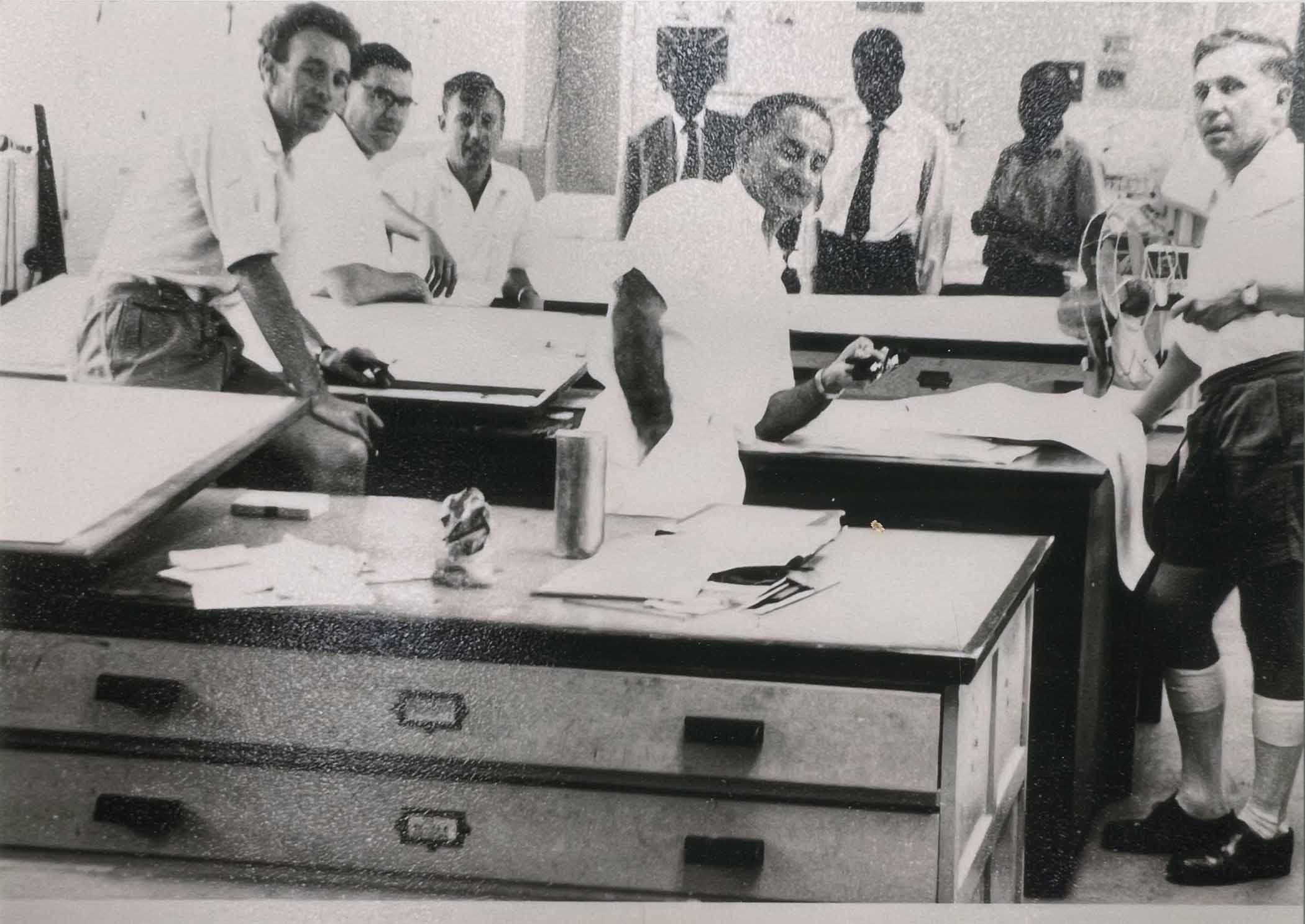 A group of men in a room with desks and papers. One man is pointing.