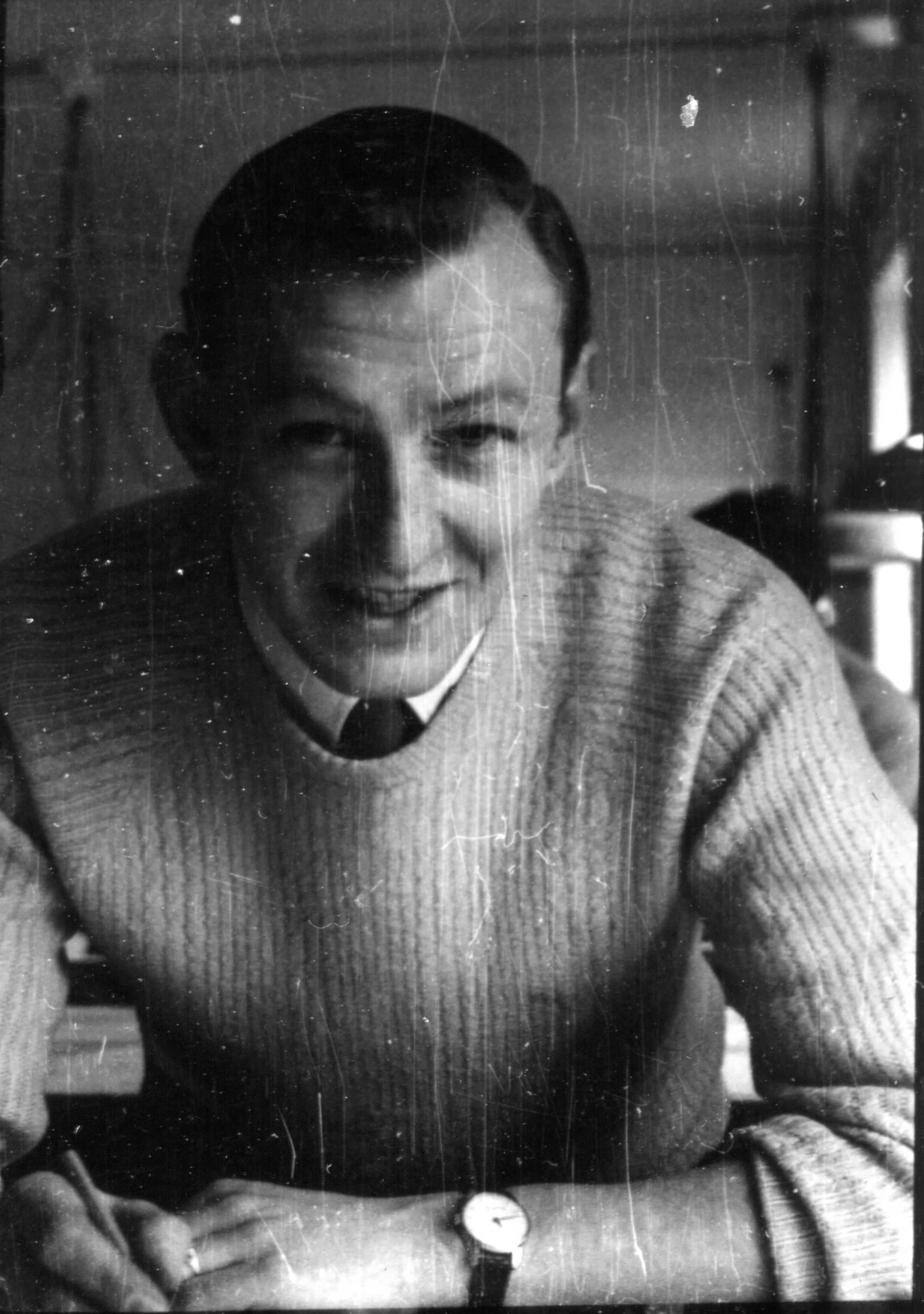 Man in a light sweater and tie looking at the camera, holding a pen.