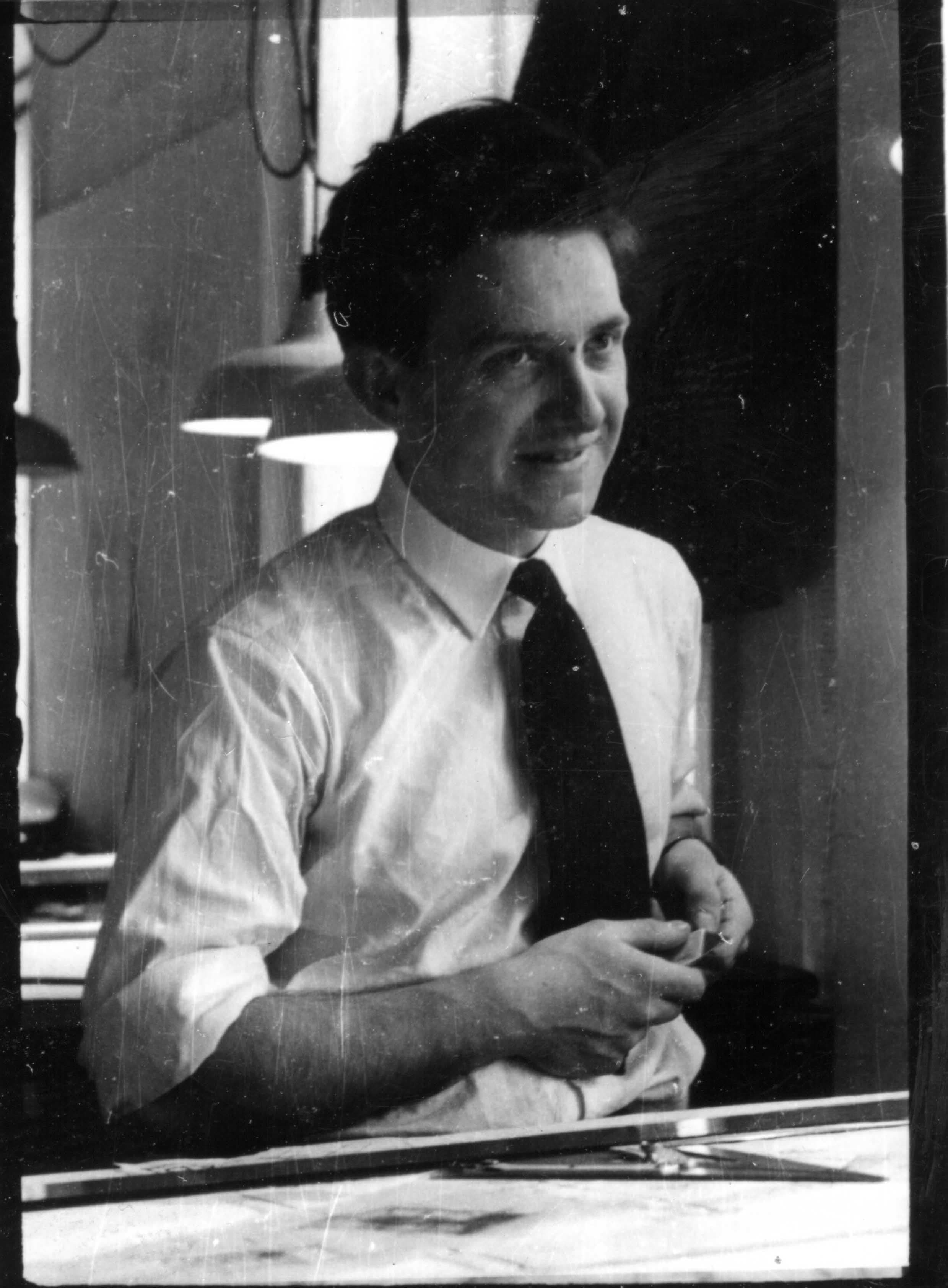 Smiling man in a white shirt and tie leaning over a desk, holding small objects.
