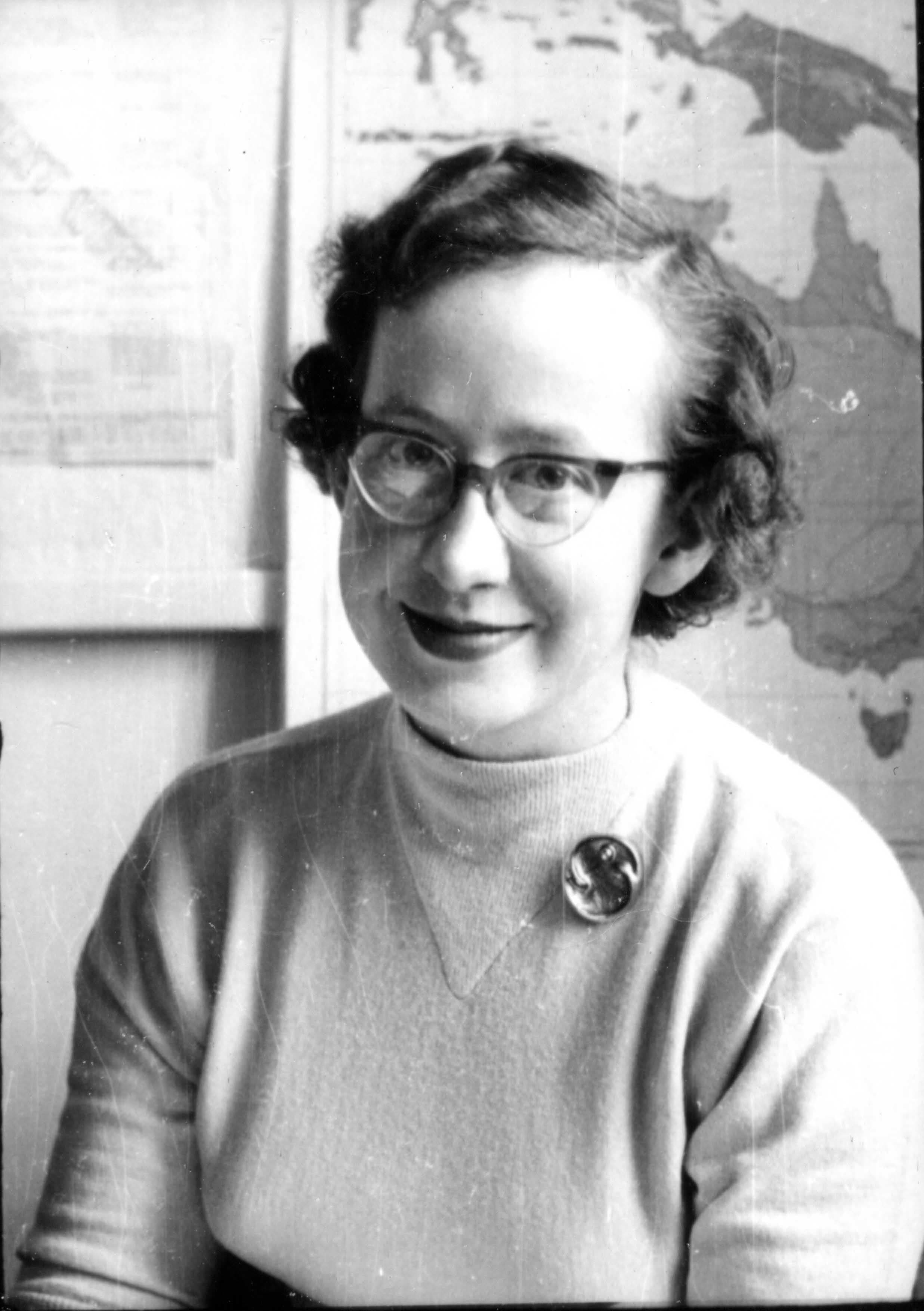 Smiling woman with glasses, light sweater, and brooch, with a world map behind her.