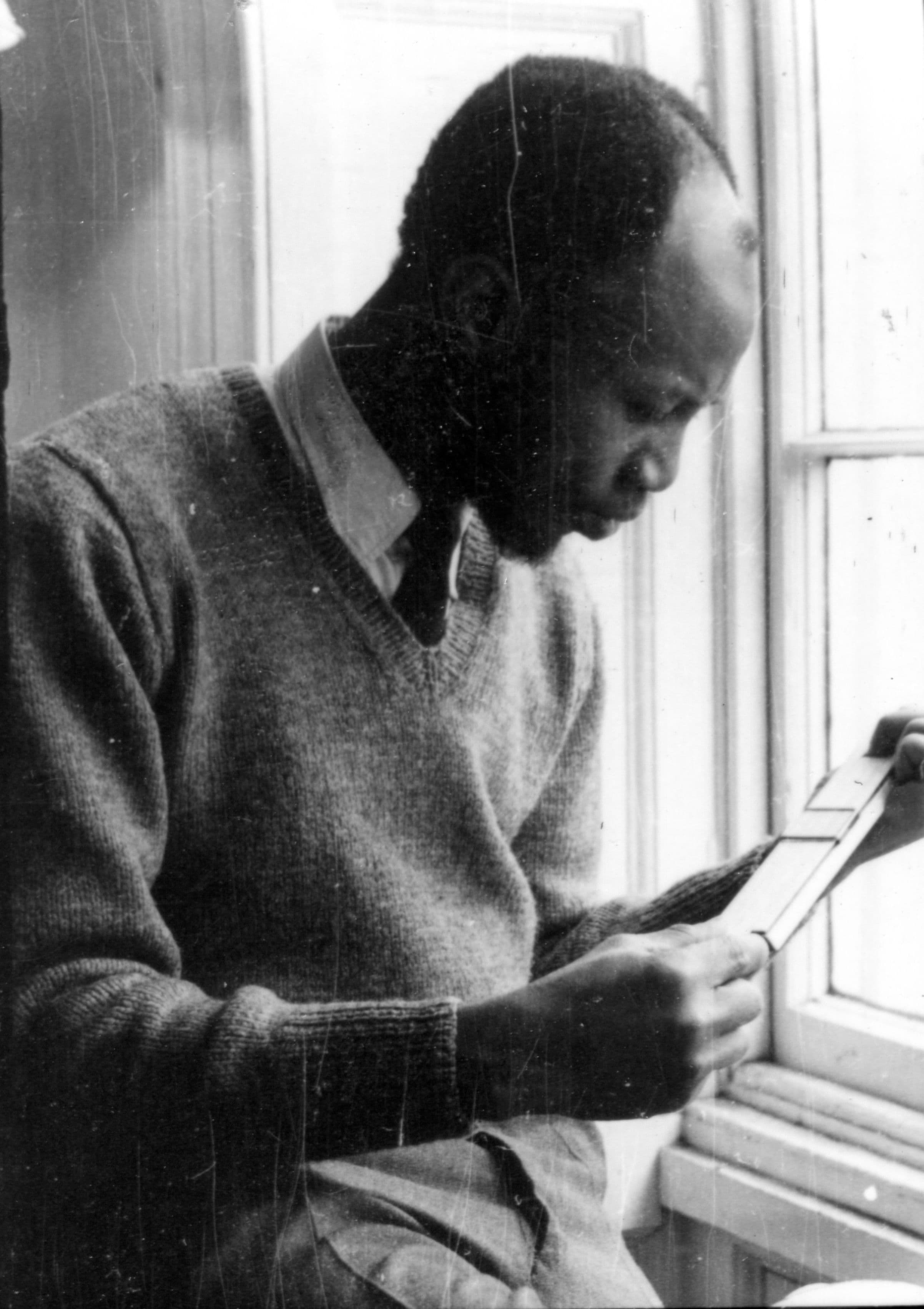 Man wearing a sweater and tie examines a document near a window.
