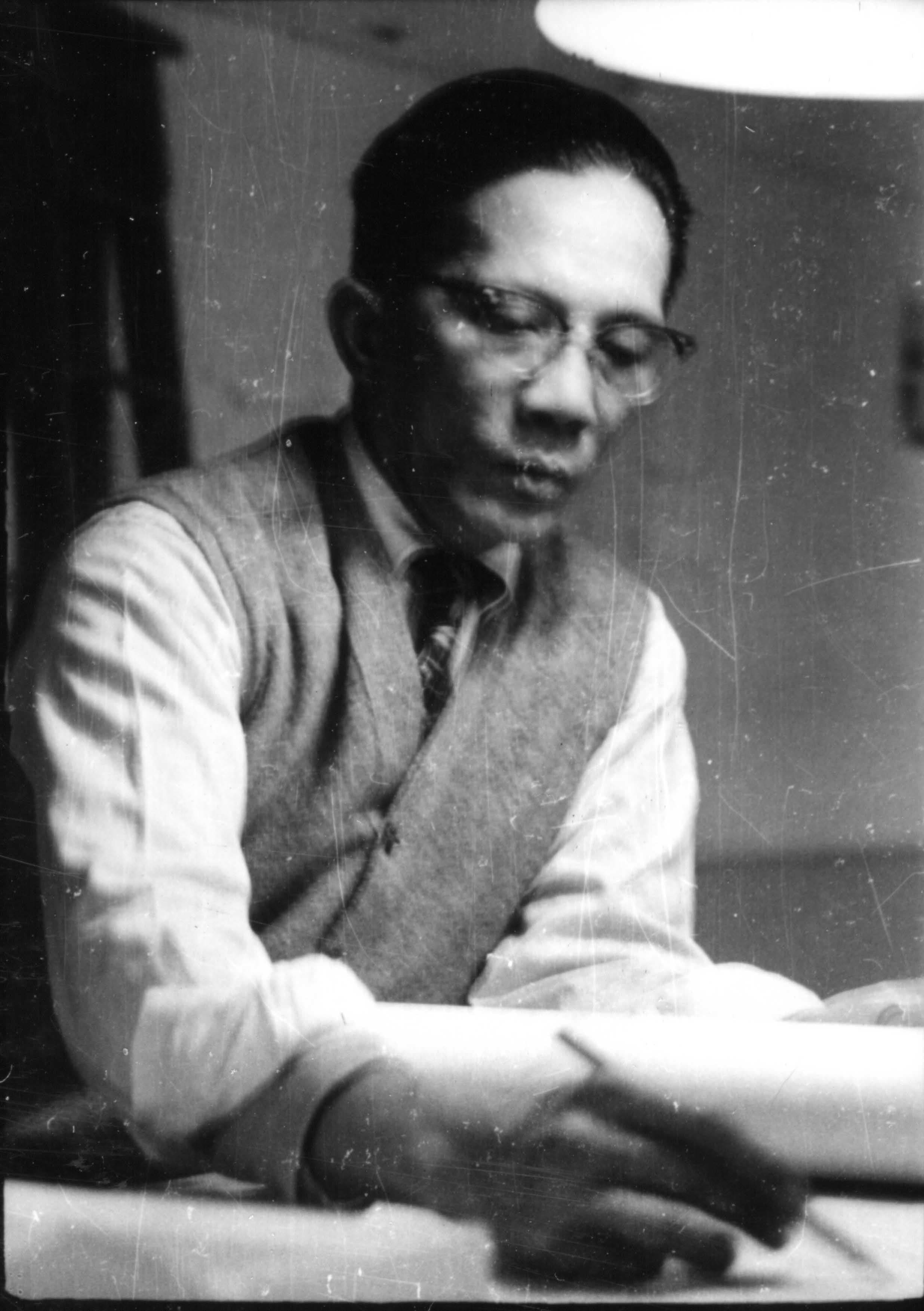 Man with glasses, white shirt, vest, and bow tie writing at a desk.