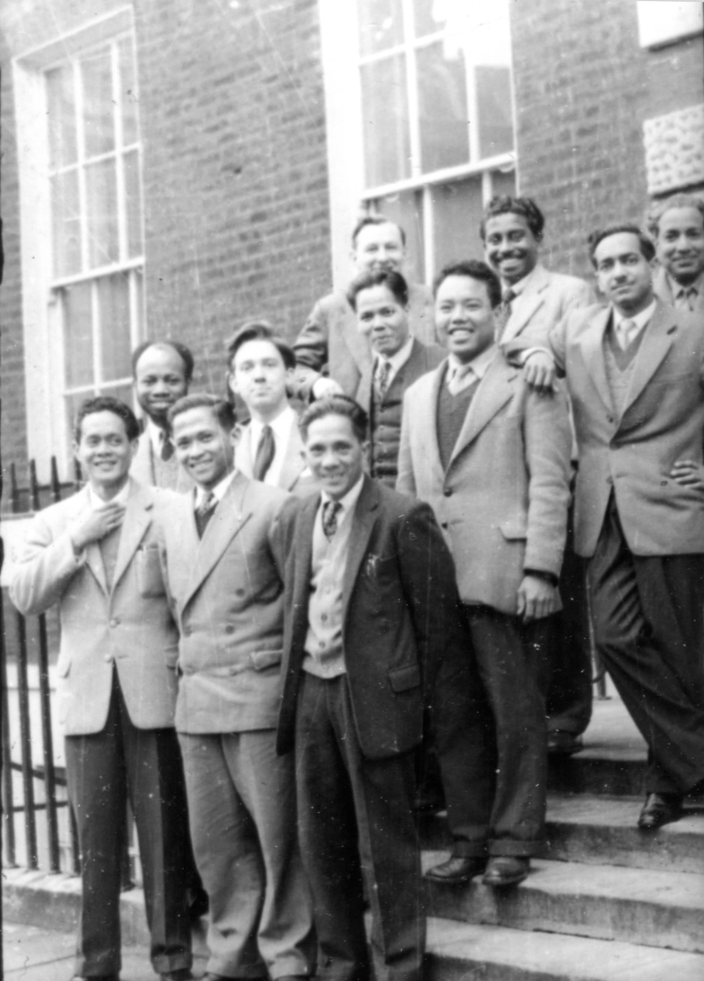 Group of eleven men in suits and ties standing on steps outside a brick building.