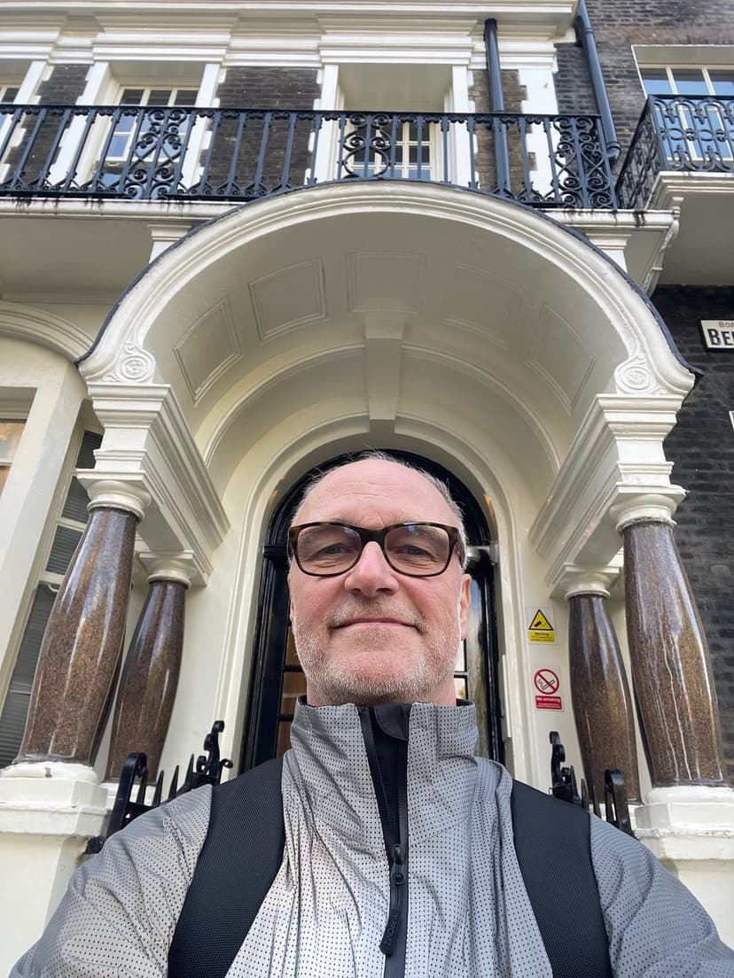 A man with glasses and a gray jacket stands in front of an ornate building entrance.