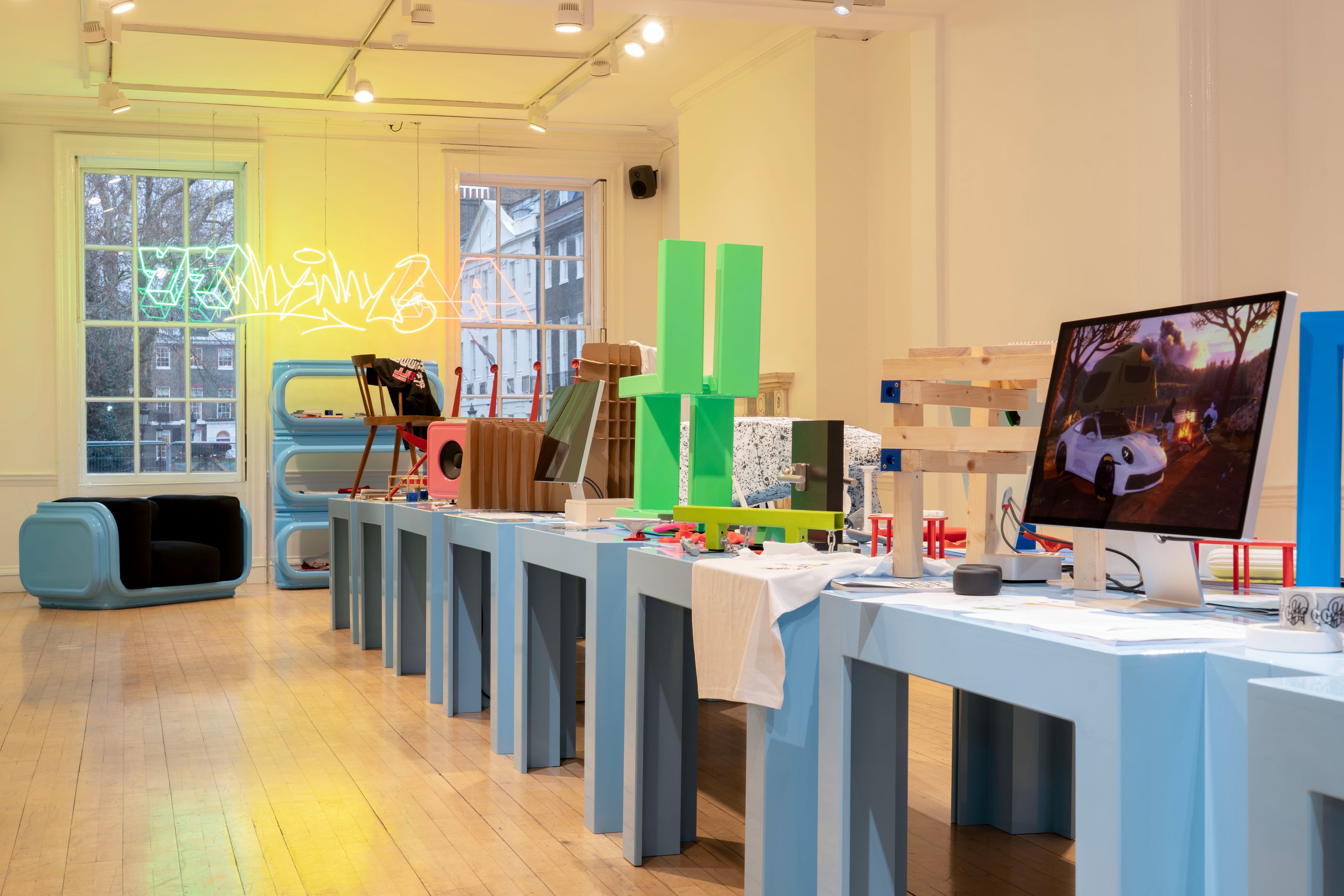 Objects on a long blue table in a gallery space with a coloured neon sign mounted on the far wall.