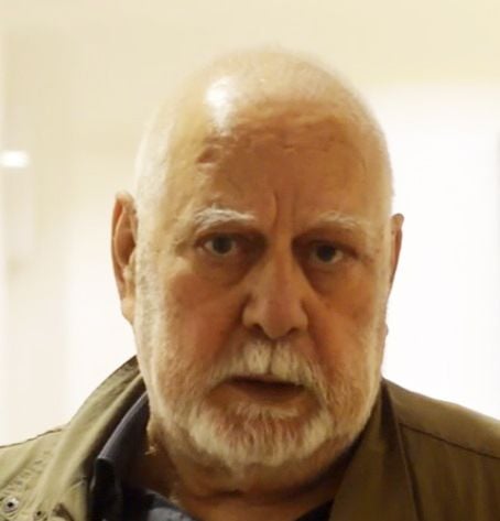 A bald older man with a white beard and mustache looks thoughtfully at the camera.