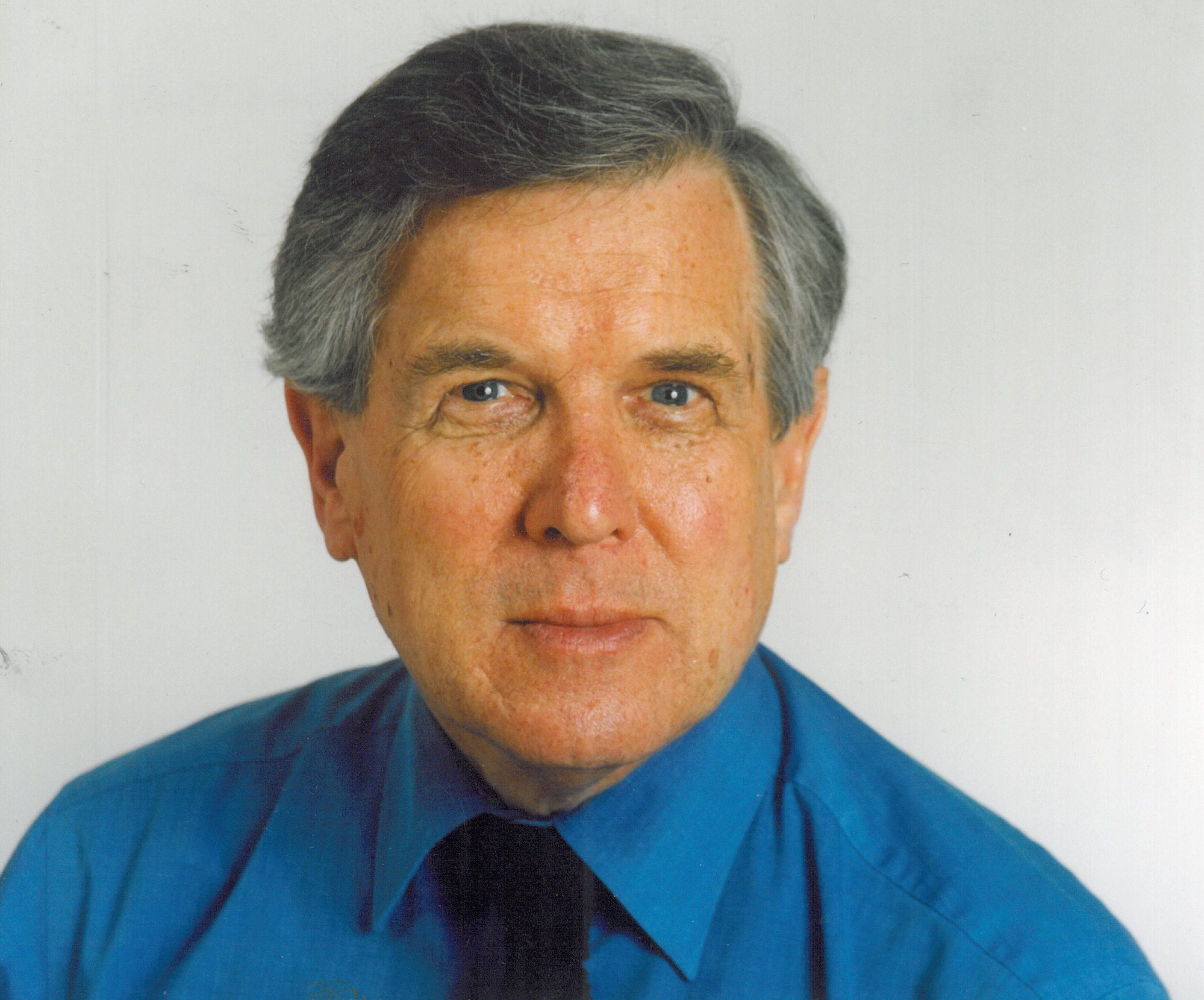 An older Caucasian man with grey hair, wearing a blue collared shirt and dark tie, looks directly at the camera.