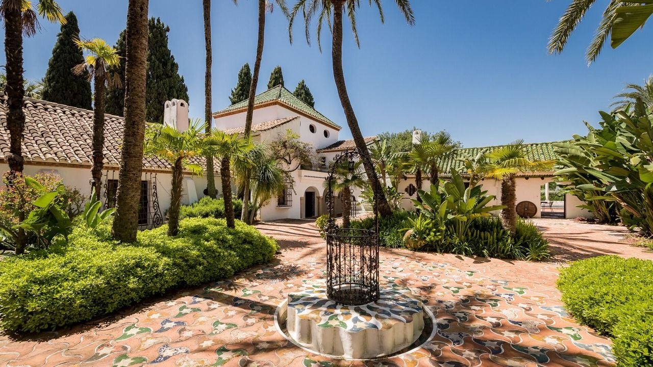 A sunny Mediterranean courtyard with a white building, tall palm trees, and a central water feature.