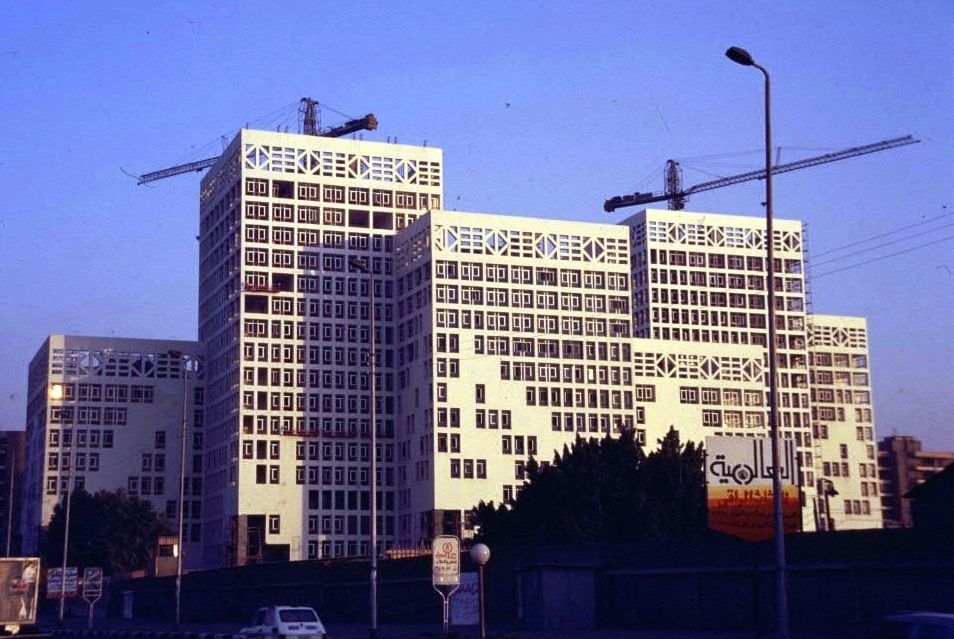 Modern multi-story white building complex with construction cranes under a clear sky.
