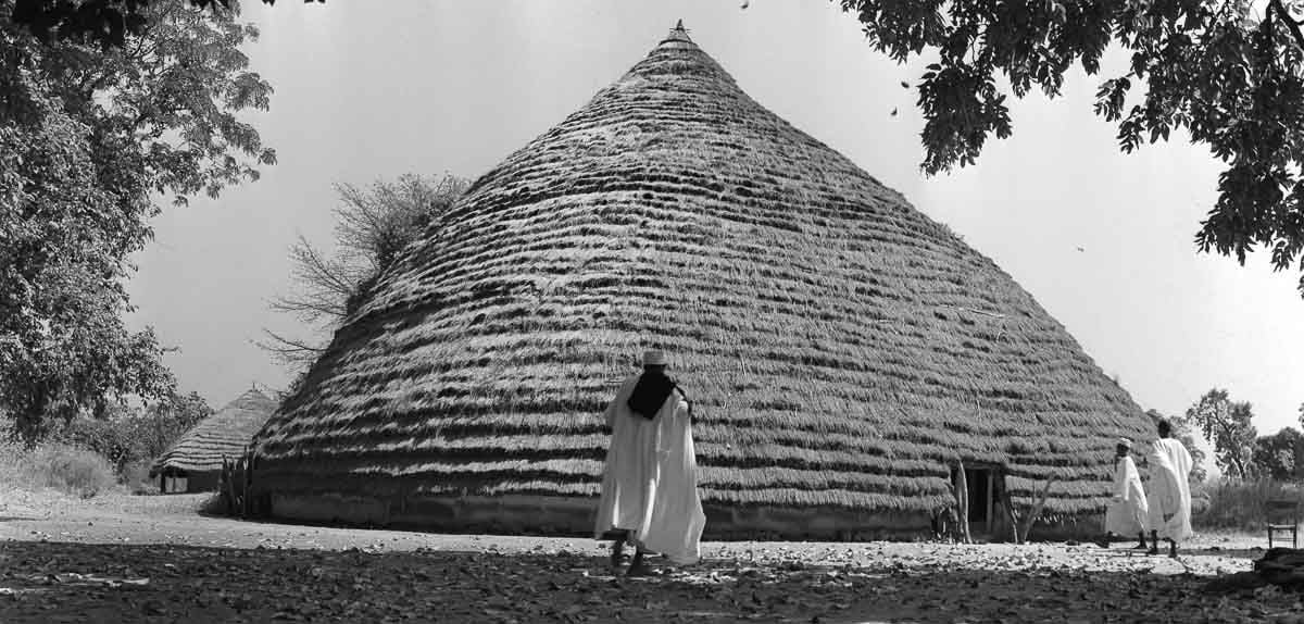 A large, conical thatched building with a doorway and a few people standing nearby.
