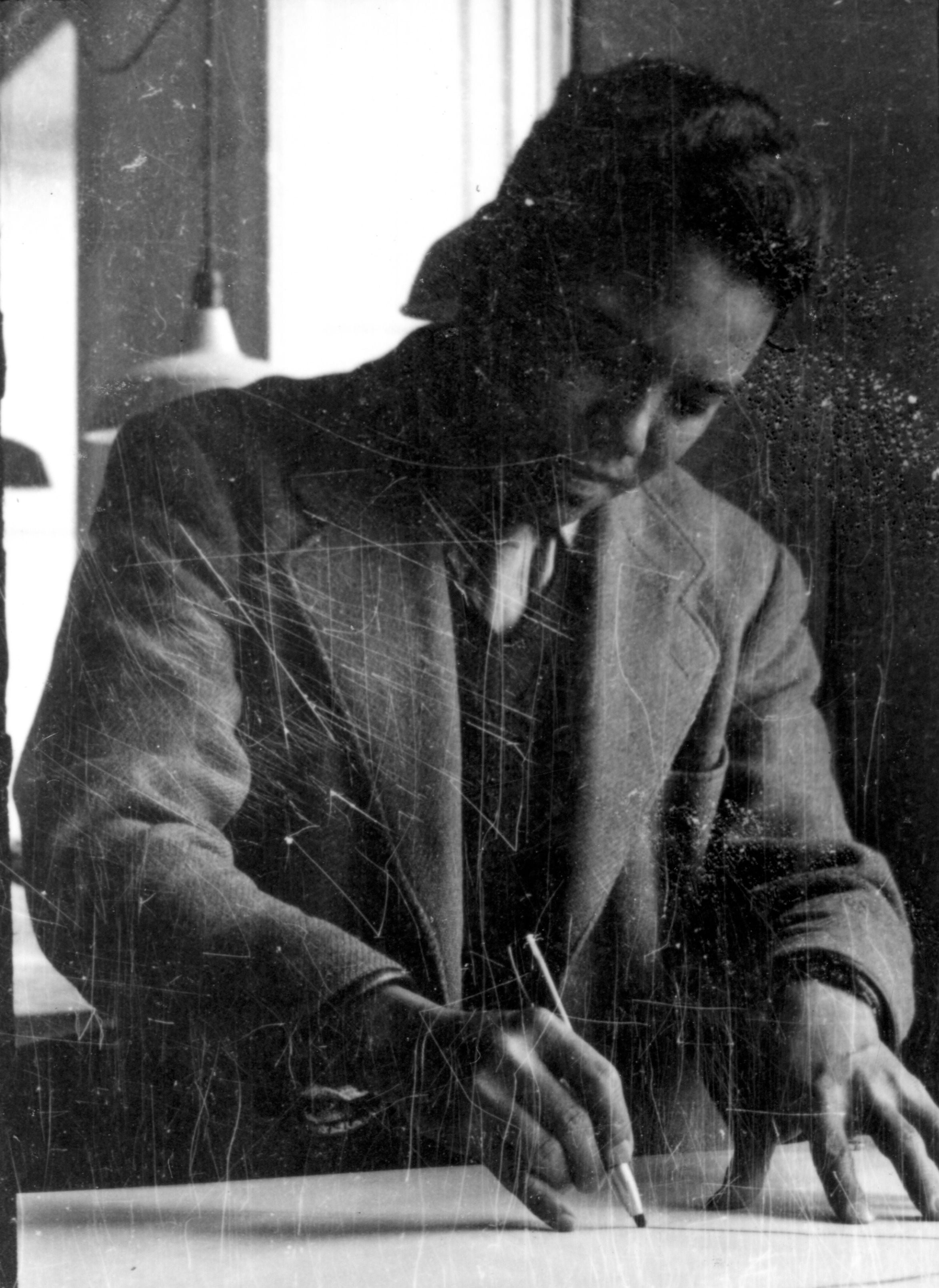 A man in a suit jacket leans over a table, drawing with a pencil.
