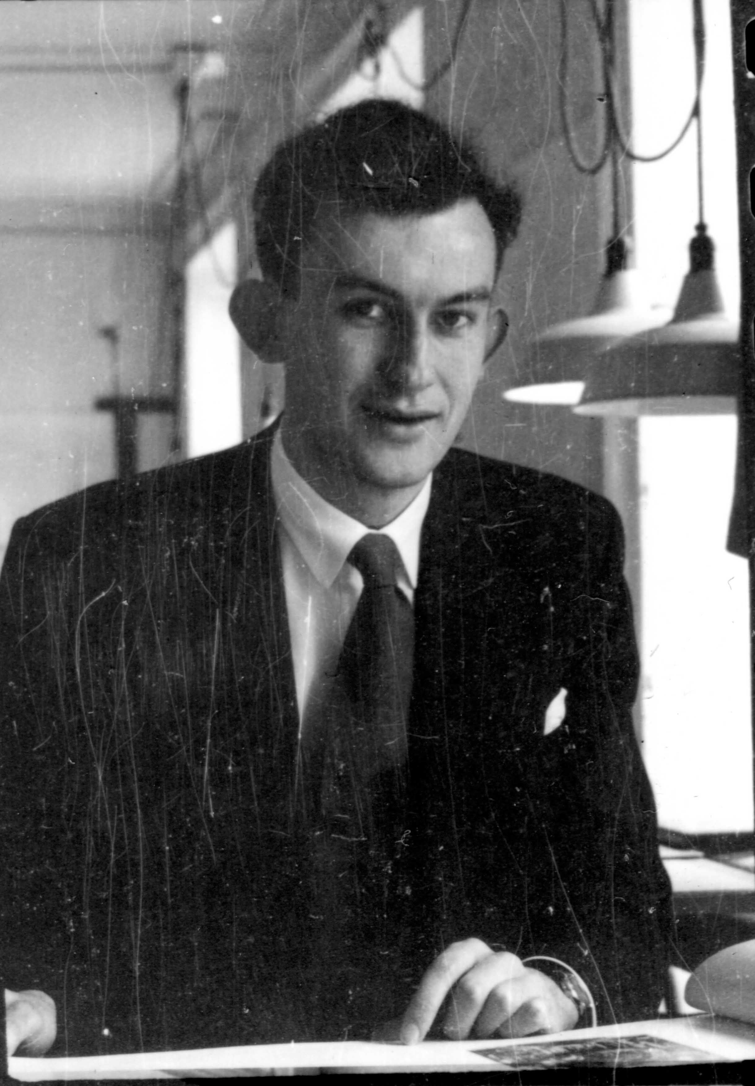 Man in a dark suit and tie looking directly at the camera while seated at a desk.