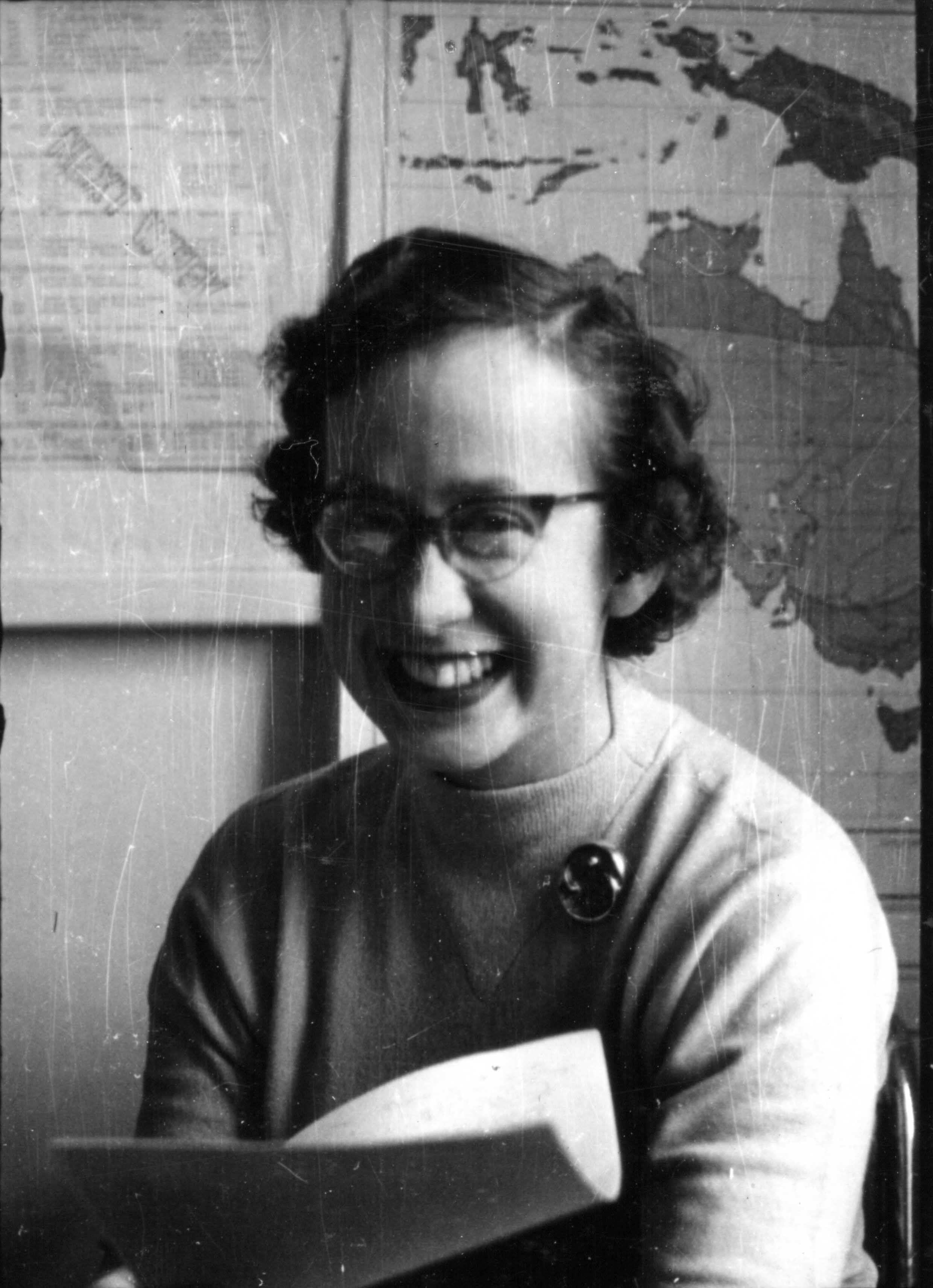 Smiling woman with glasses, light sweater, and brooch, with a map in the background.