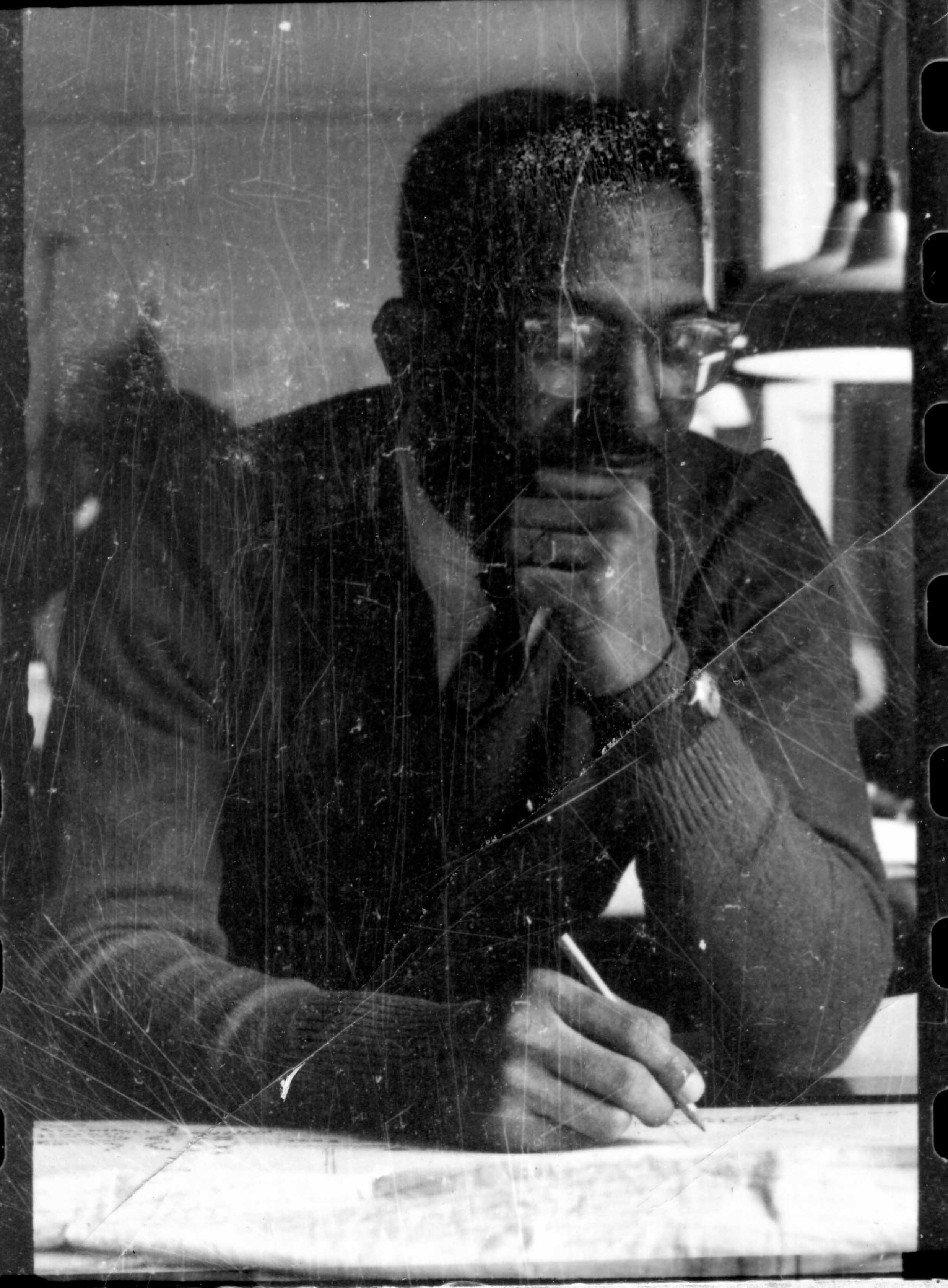 Man with beard and glasses, in a suit, writing at a desk, deep in thought.