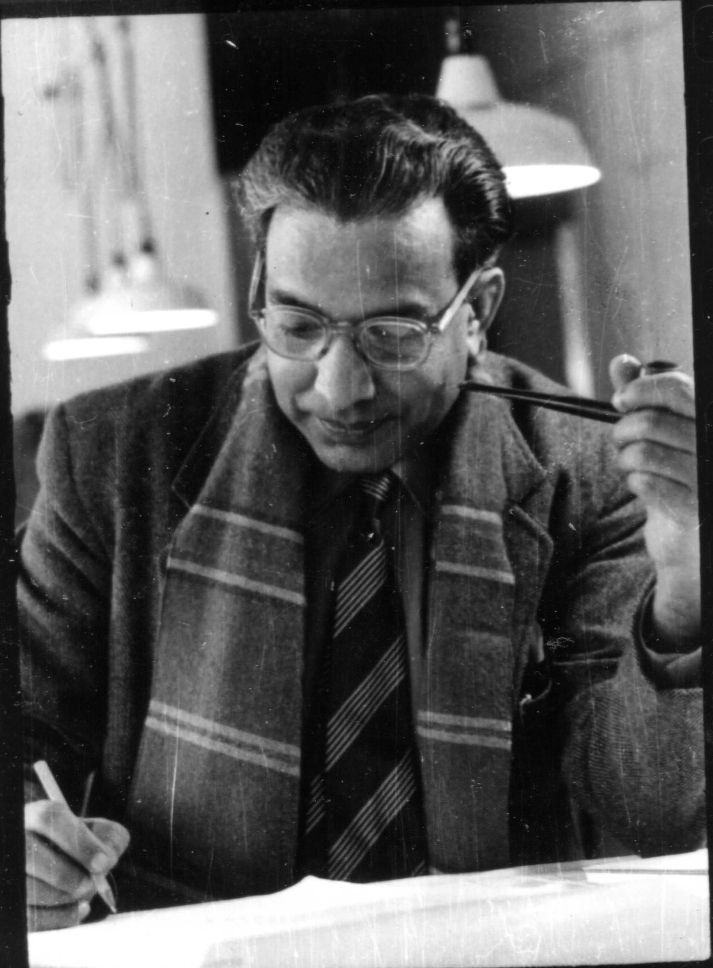 Man in glasses, suit, scarf, and tie, smoking a pipe while writing at a desk.