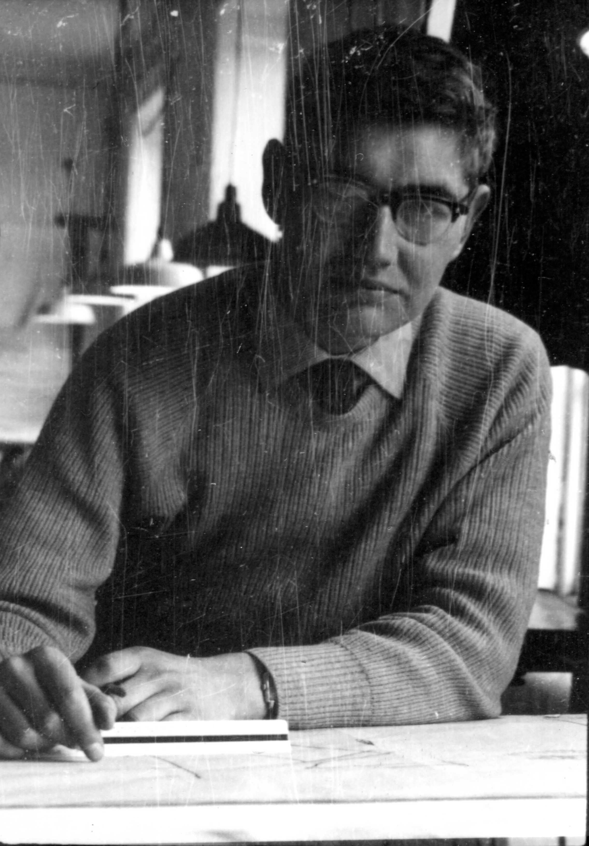 Man in glasses, sweater, and shirt, sitting at a desk looking at the viewer.