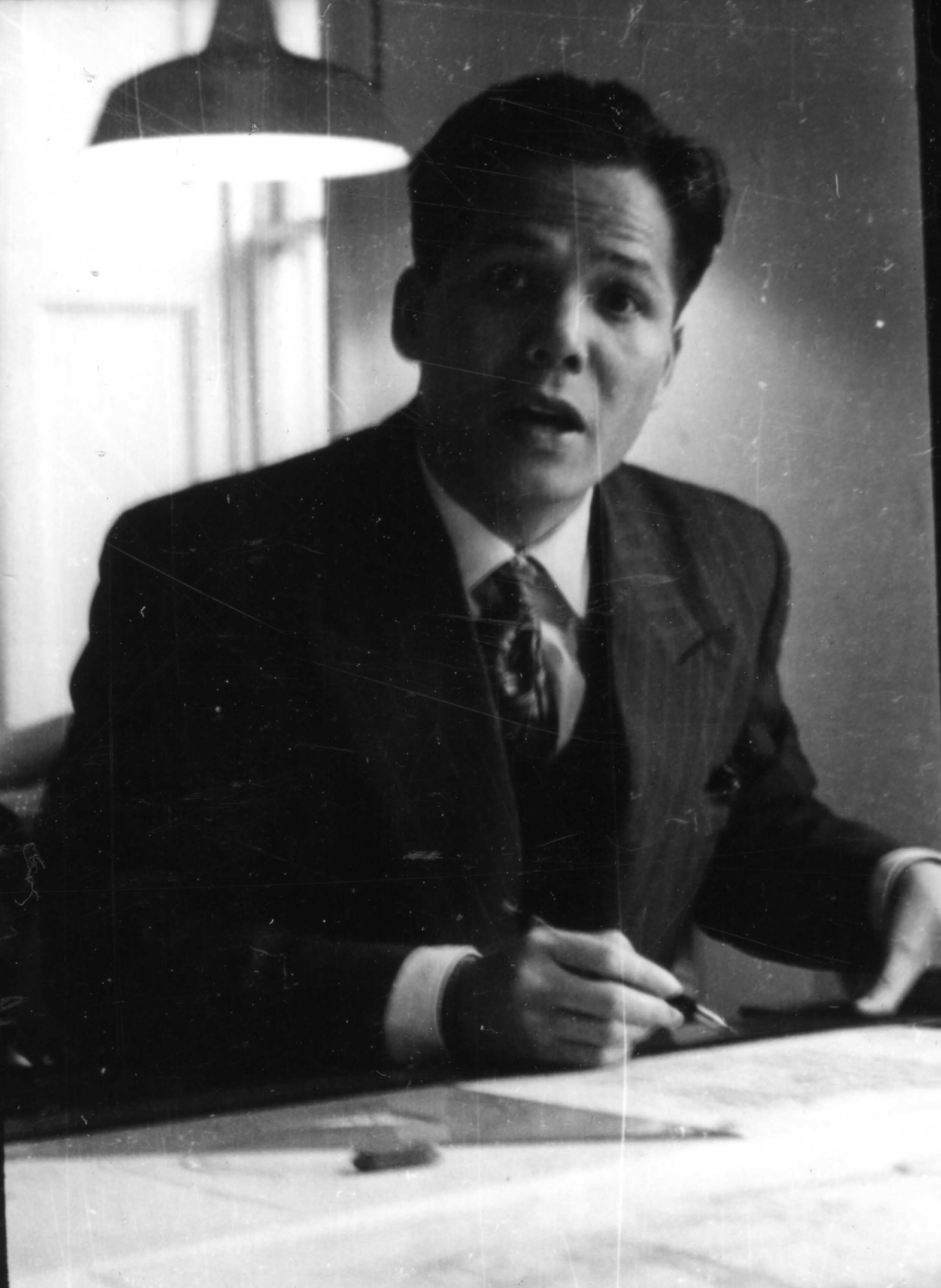 Man in a suit and tie, sitting at a desk, looking up and speaking.