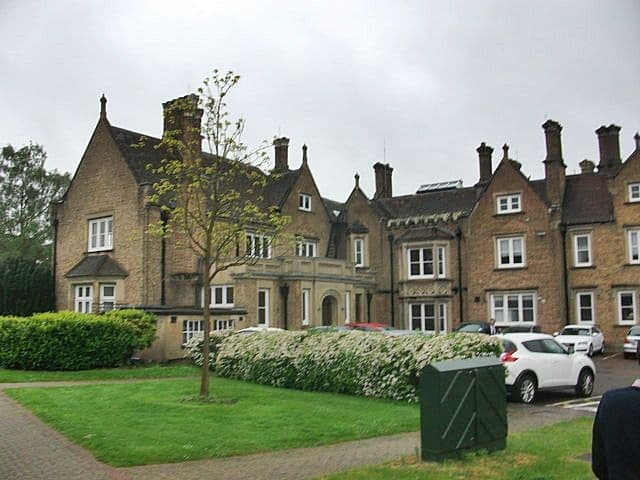 A large, elegant traditional mansion with a green lawn and parked cars on a cloudy day.