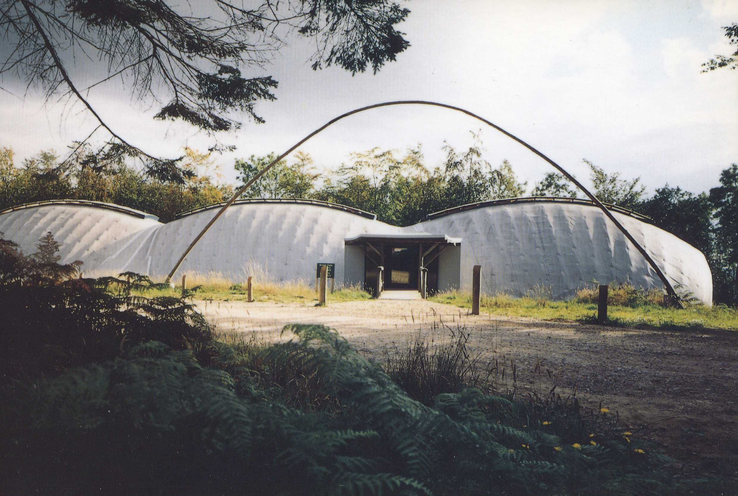 Curving, white, tent-like structure with a prominent arch over the entrance, surrounded by trees and foliage.