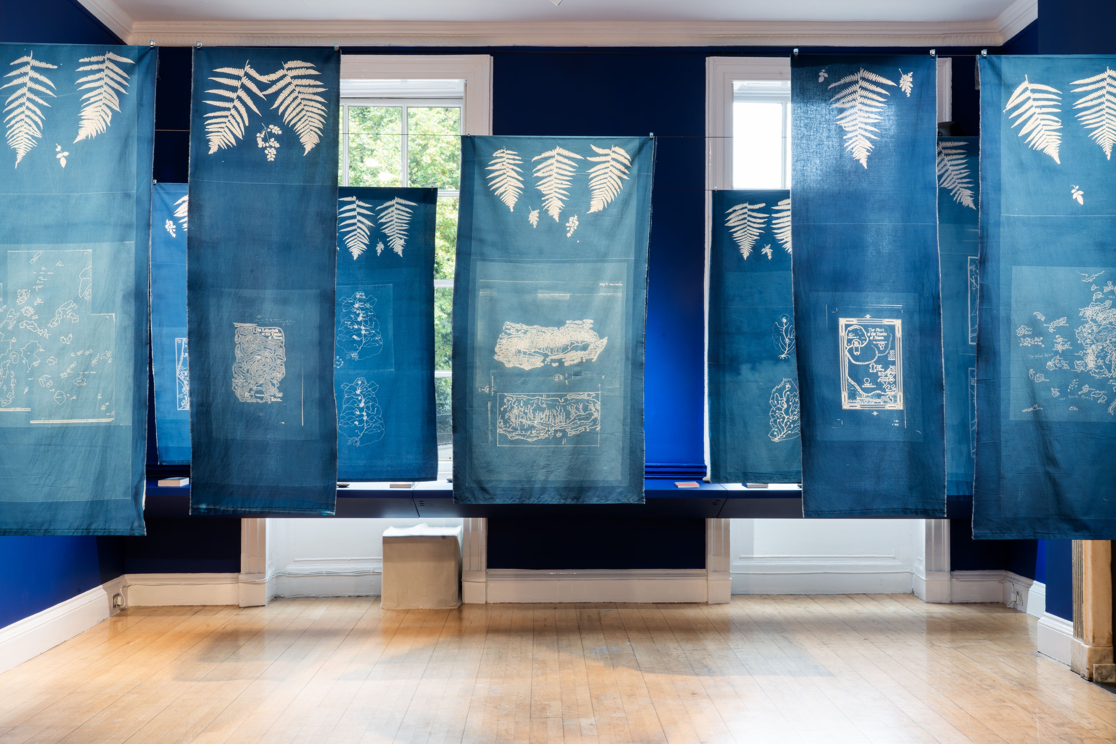 Cyanotype prints of maps and leaves on fabric against a gallery painted ultramarine blue.