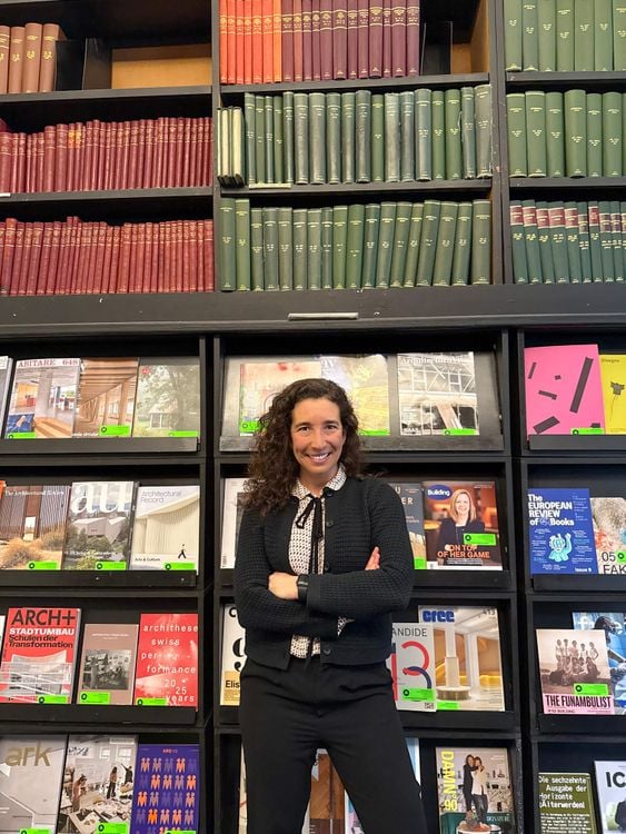 A woman with curly hair smiles, arms crossed, in front of a wall of books.