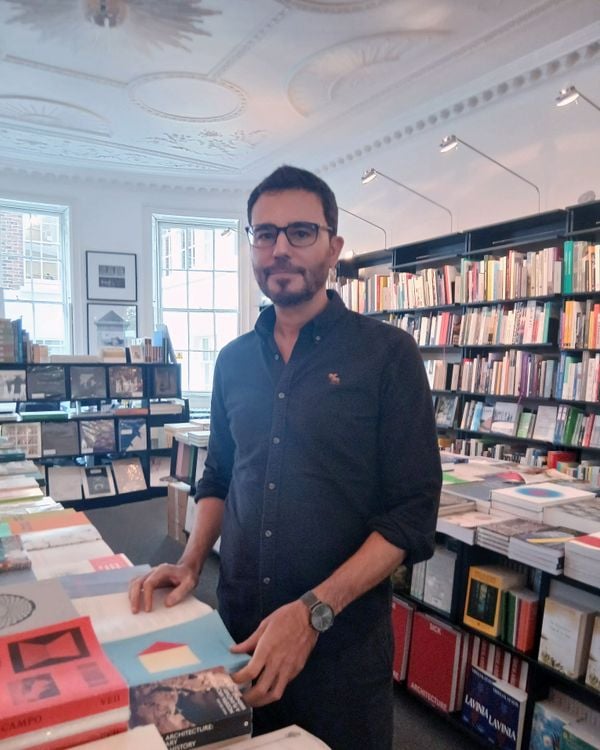 A man with short dark hair, glasses, and a beard wears a dark shirt in a bookstore.
